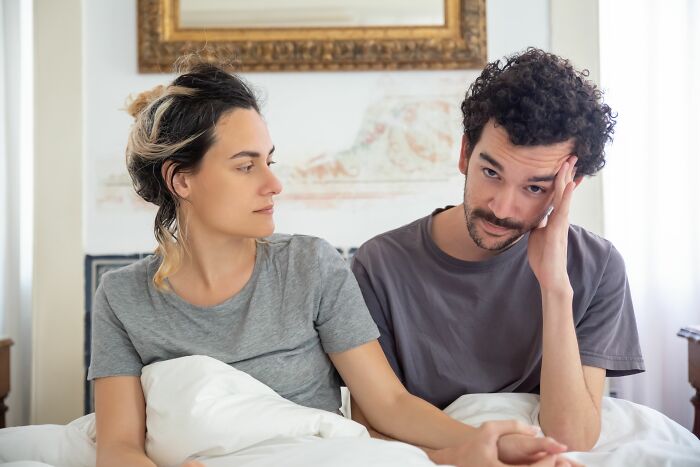 An identical twin couple in bed, one looking worried and the other serious, displaying pure chaos.