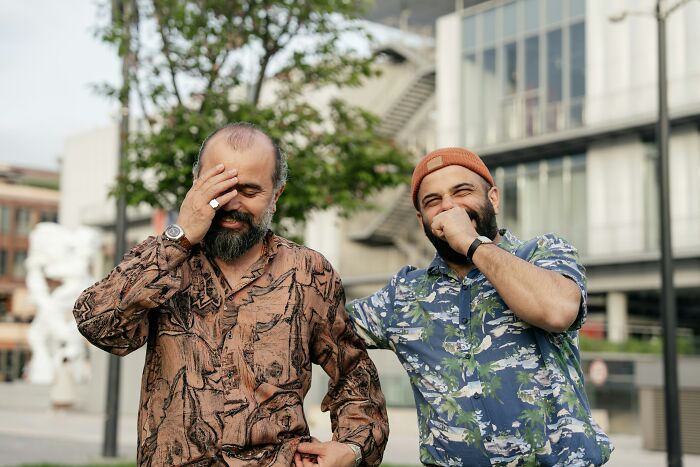 Two identical twin men laughing and covering their faces while standing outdoors, showing pure chaos.