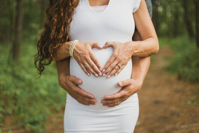A pregnant woman in white, hands forming a heart shape over her belly, held by a man's arms. A moment of calm amidst the pure chaos of identical twin parenthood.