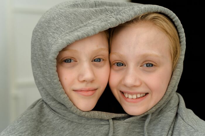 Close-up of two identical twin girls with their heads touching, both wearing a grey hoodie, illustrating the chaos of being a twin.