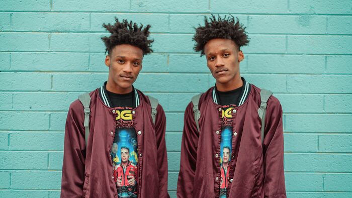 Two identical twin brothers with matching outfits stand against a blue brick wall, showing the chaos of being a twin.
