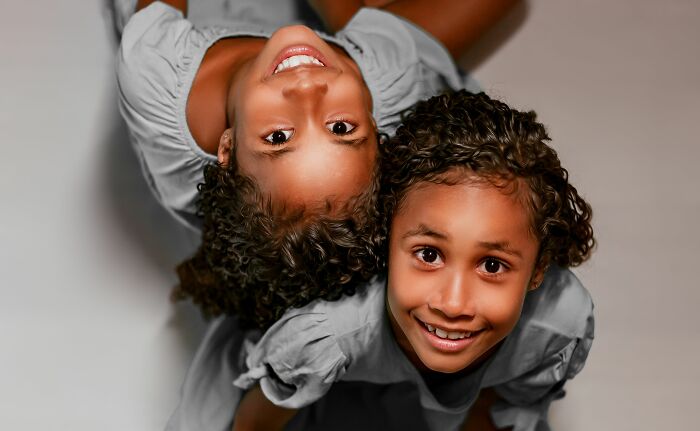 Two young, smiling identical twin girls with curly hair look up at the camera, their heads close together.