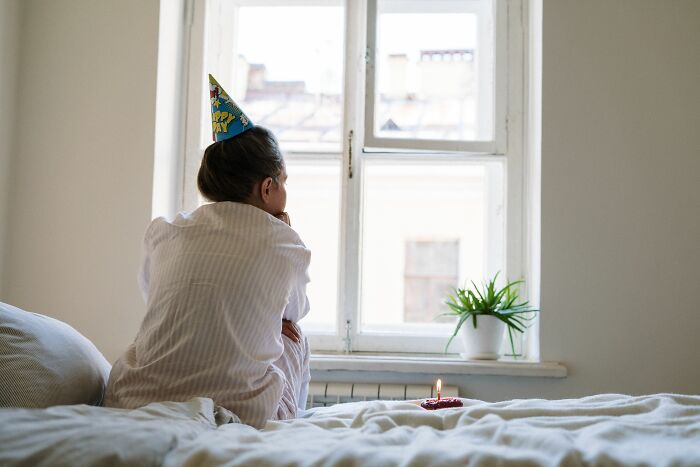 A young child in a party hat sits on a bed, looking out a window with a lit candle nearby, creating a sense of isolation.