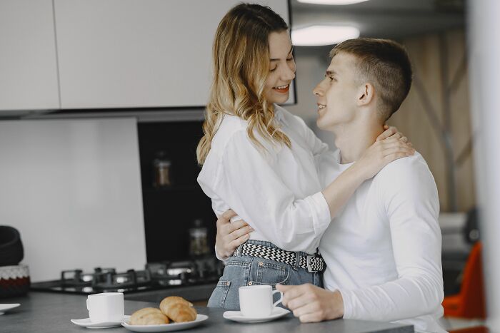 A couple embraces in a modern kitchen, with the woman looking at the man, suggesting a moment of pure chaos.