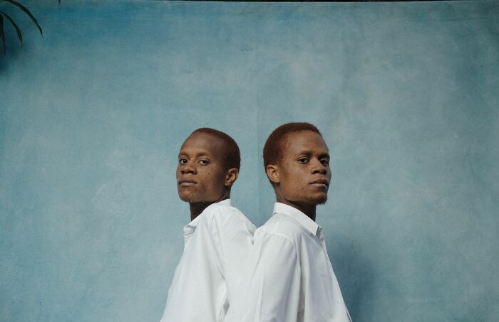 Two young men, identical twins, in white shirts stand back-to-back, looking at the camera with serious expressions.