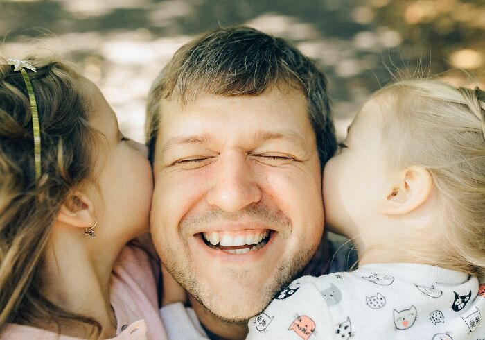 A happy father with his eyes closed, smiling as his two identical twin daughters kiss him on each cheek, showing pure chaos.