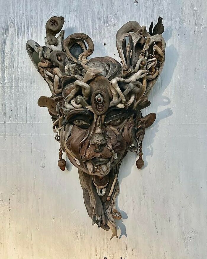Detailed wood sculpture of a face with elaborate hair made from collected natural pieces, hanging on a white wall.