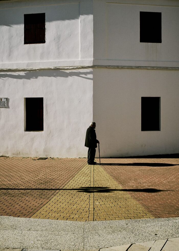 An extraordinary moment in a street photo shows an old man with a cane, casting a long shadow on a checkered path.