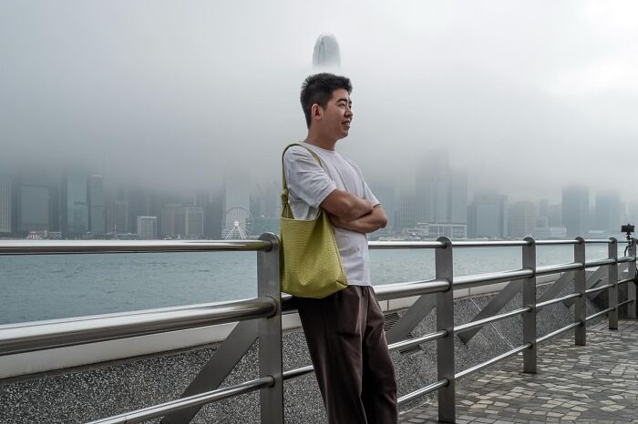Street coincidence: A man leaning on a railing with a distant skyscraper resembling a hat, by Edas Wong.