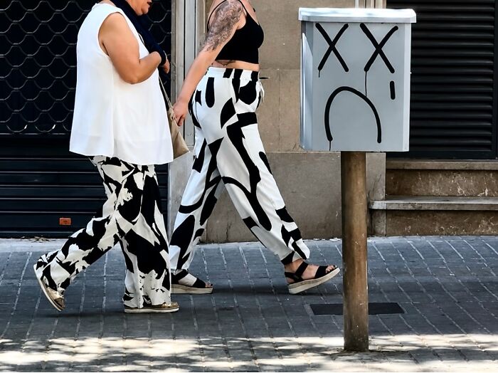 Two women with matching black and white patterned pants walk past a sad-faced mailbox in Barcelona, a perfectly timed coincidence.