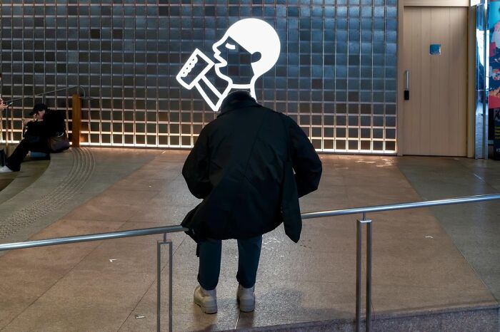 A person sitting on a rail aligns perfectly with a light-up figure drinking from a mug, an unbelievable street coincidence.