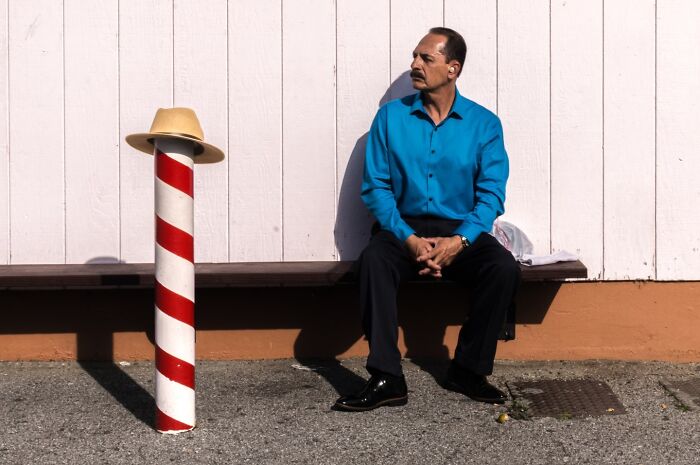 A photographer captures humor and chaos of a man sitting on a bench next to a striped pole in everyday life.