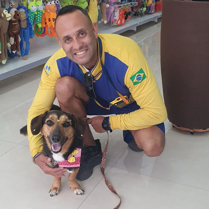 A kind mailman crouches, smiling with a small dog wearing a decorative collar, on a leash indoors.