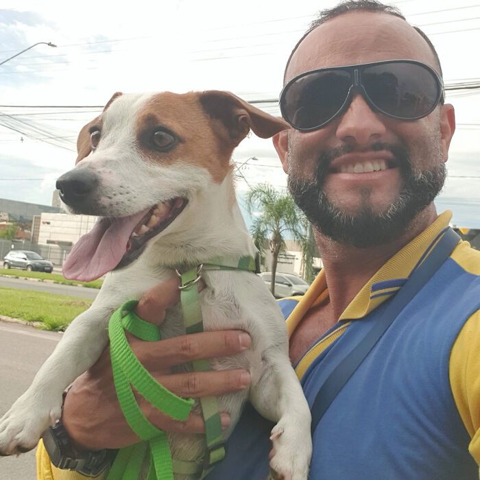 A mailman in sunglasses smiles while holding a happy dog with a green leash, befriending dogs during his deliveries.