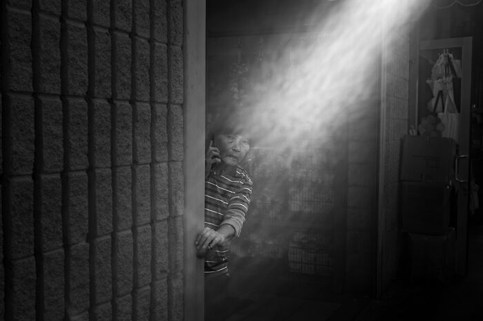 A photographer captures humor in everyday life; an elderly person on the phone peeks from a doorway in dramatic light.
