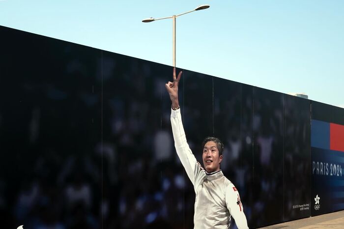 A man in fencing attire with his arm raised, perfectly aligning his finger with a lamppost, a street coincidence.
