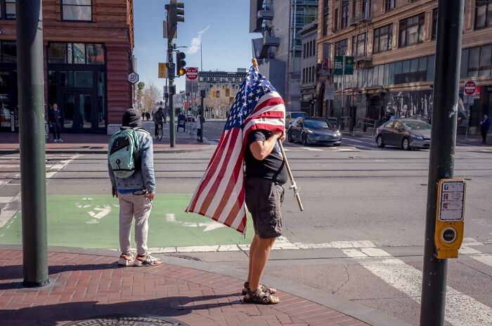 A street photographer finds humor in everyday life; a person holding an American flag obscuring their face on a city street.
