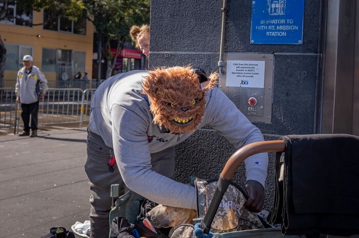 Everyday life humor captured by a photographer; a person in a monster mask bends over a stroller on a city sidewalk.