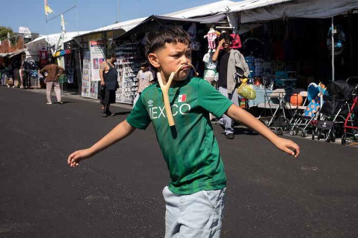 A photographer's shot of humor in everyday life; a young boy in a Mexico shirt holds a slingshot in his mouth on a street.