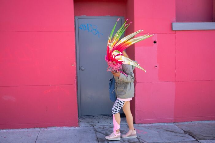 Humor in everyday life by a photographer; a person in a colorful, feathered mask stands against a bright pink wall.