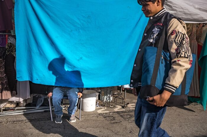 A photographer captures humor in everyday life: a man sits behind a blue tarp, his shadow visible.