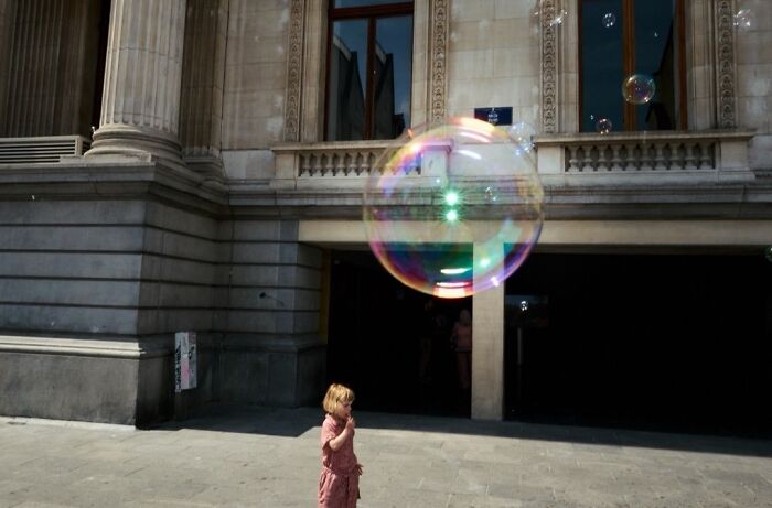 A young child watches a large, colorful bubble float by, a perfectly timed street photo with an extraordinary moment.