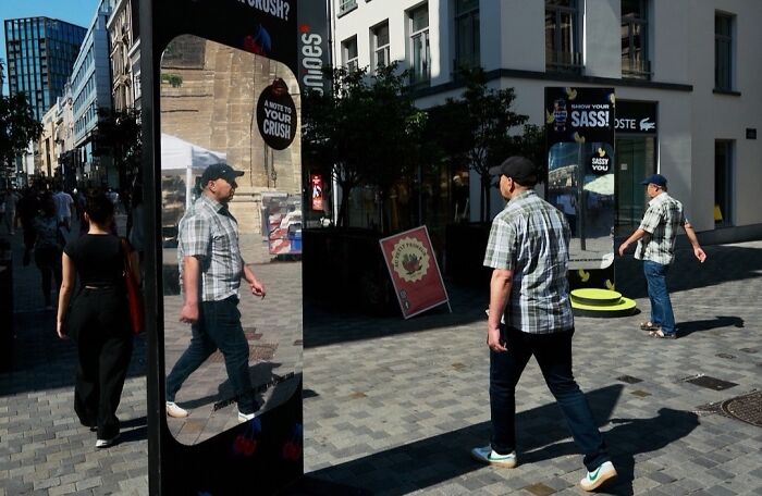 A man walks past a mirrored pillar reflecting his image, a perfectly timed street photo with extraordinary moments.