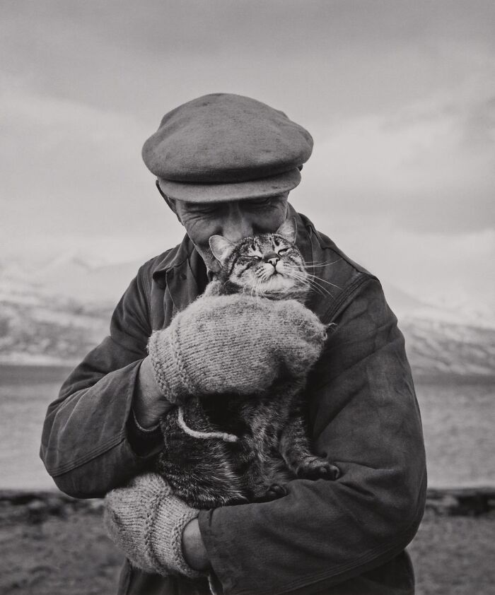Historical photos: An older man wearing a cap and mittens, affectionately holding a cat against a scenic backdrop.