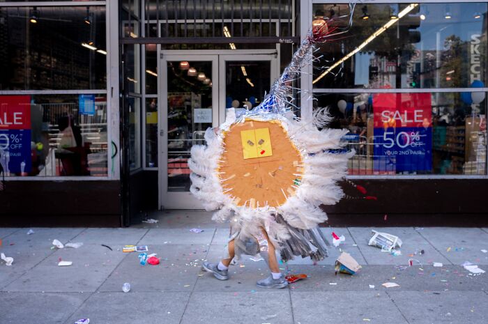 A photographer captures humor and chaos in everyday life: a person in a large, feathered costume on a littered sidewalk.