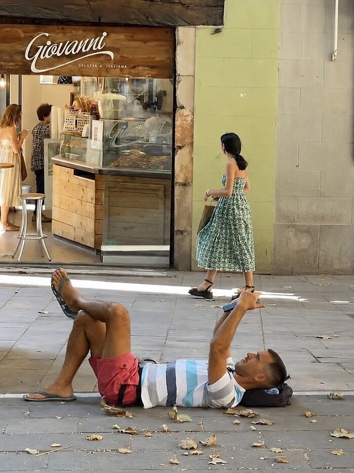 A man lies on the ground checking his phone, a woman walks by an ice cream shop in Barcelona, a perfectly timed coincidence.
