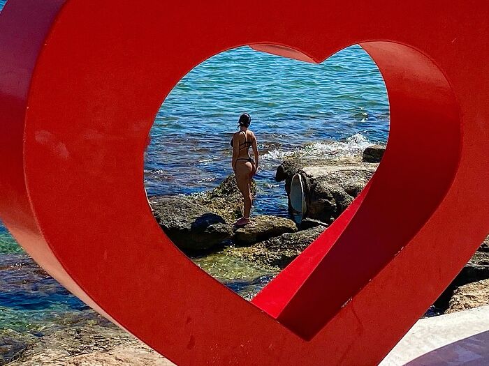 A woman stands by the sea, viewed through a large red heart sculpture, a perfectly timed coincidence in Barcelona.