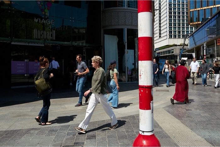A perfectly timed street photo of pedestrians walking on a city street, with a red and white pole in the foreground.