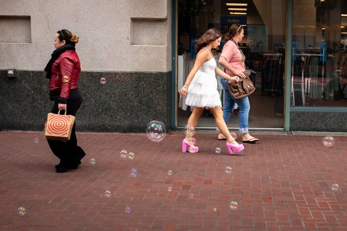 Photographer captures everyday life humor: a woman in pink platform shoes walks past bubbles on a city sidewalk.