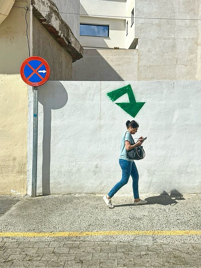A photographer captures a perfectly timed coincidence of a woman walking under a green arrow pointing down, while a no-entry sign is nearby in Barcelona.