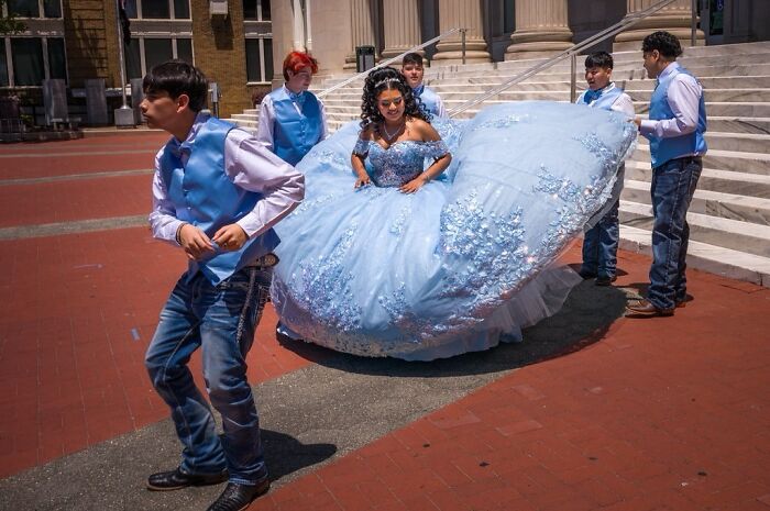 Young men help a girl in a large blue gown navigate a public space, illustrating humor and chaos in everyday life.