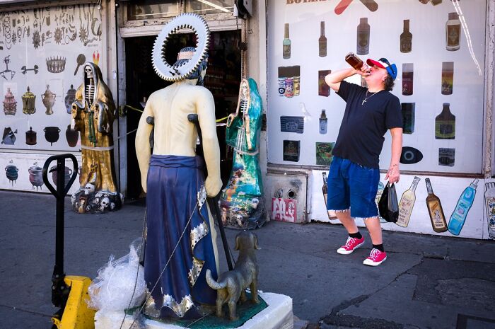 A man drinks from a bottle next to a statue of a saint on a sidewalk, showcasing humor and chaos in everyday life.