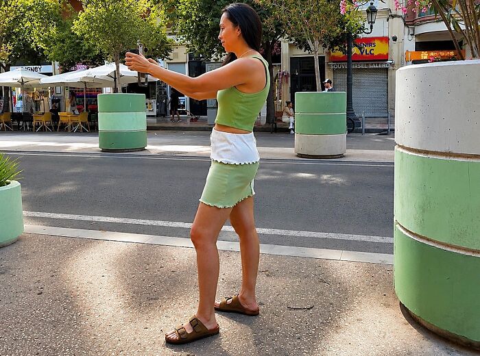 A woman in a green top and skirt taking a selfie on a sunny street, capturing perfectly timed coincidences.