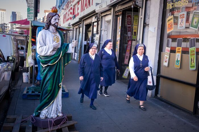 Three nuns walk past a statue of a saint on a city street, capturing humor and chaos in everyday life.