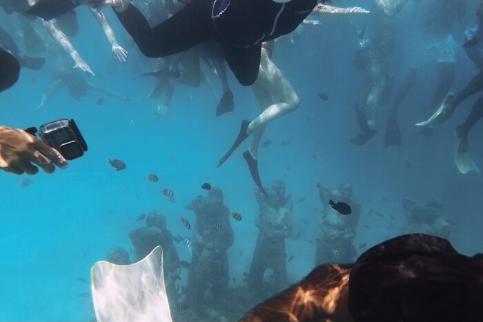 A perfectly timed street photo of people swimming underwater with fish and submerged statues.