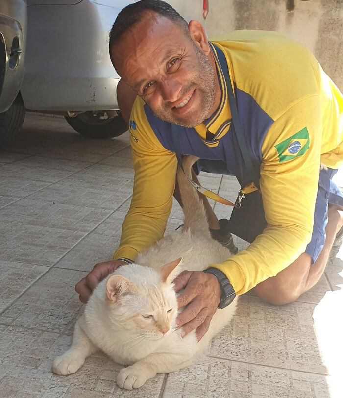 A kind mailman kneels and gently pets a light-colored cat on a tiled patio during his deliveries.