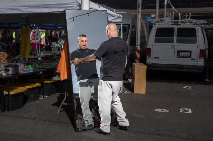 A man with tattoos poses in front of a mirror at an outdoor market, reflecting humor and chaos in everyday life.