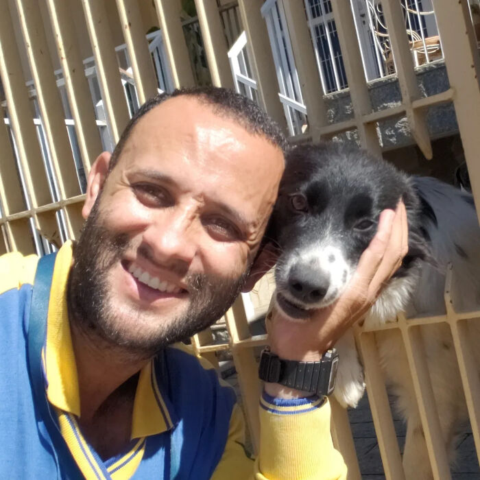 A kind mailman happily poses for a selfie, gently petting a small dog that peeks through a gate.