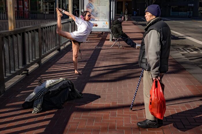 A woman doing a yoga pose on a city sidewalk next to a man with a cane, depicting humor and chaos in everyday life.