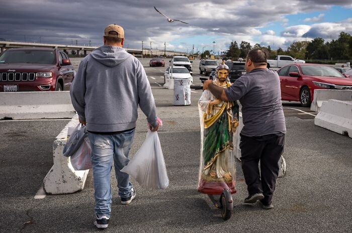 Photographer captures everyday life humor: two men transporting a large religious statue on a scooter in a parking lot.