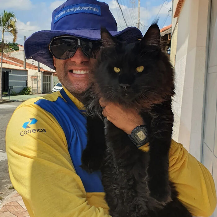 A kind mailman in a blue hat and sunglasses holds a fluffy black cat with bright yellow eyes.
