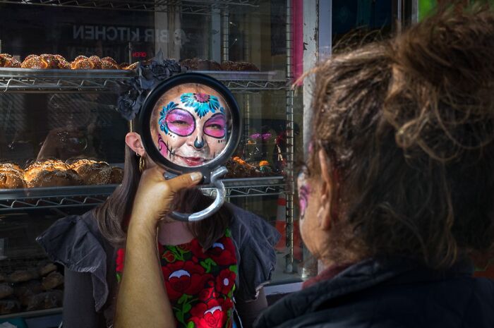 Photographer captures humor and chaos in everyday life, with a person reflecting face paint in a handheld mirror.