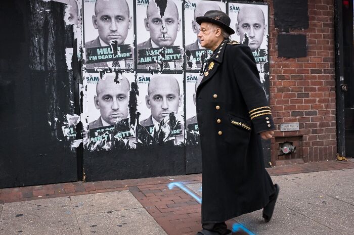 A photographer captures humor and chaos: a man in a long coat walks past a wall with multiple posters.