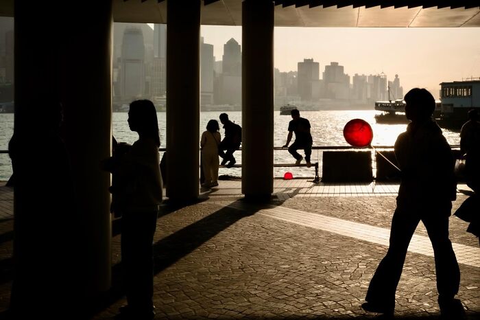 Silhouettes of people and a red balloon on a waterfront promenade with city buildings and a sunset reflection, a street coincidence.