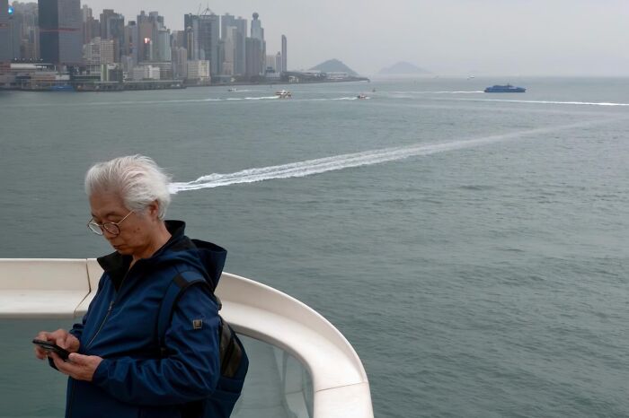 An older man with white hair looks at his phone with the Hong Kong skyline and boats on the water behind him, a street coincidence.