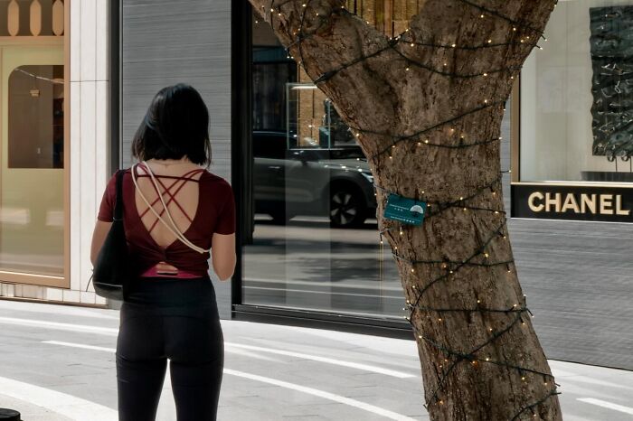 A woman in a red top with a unique back design walks past a tree wrapped in lights, a street coincidence.